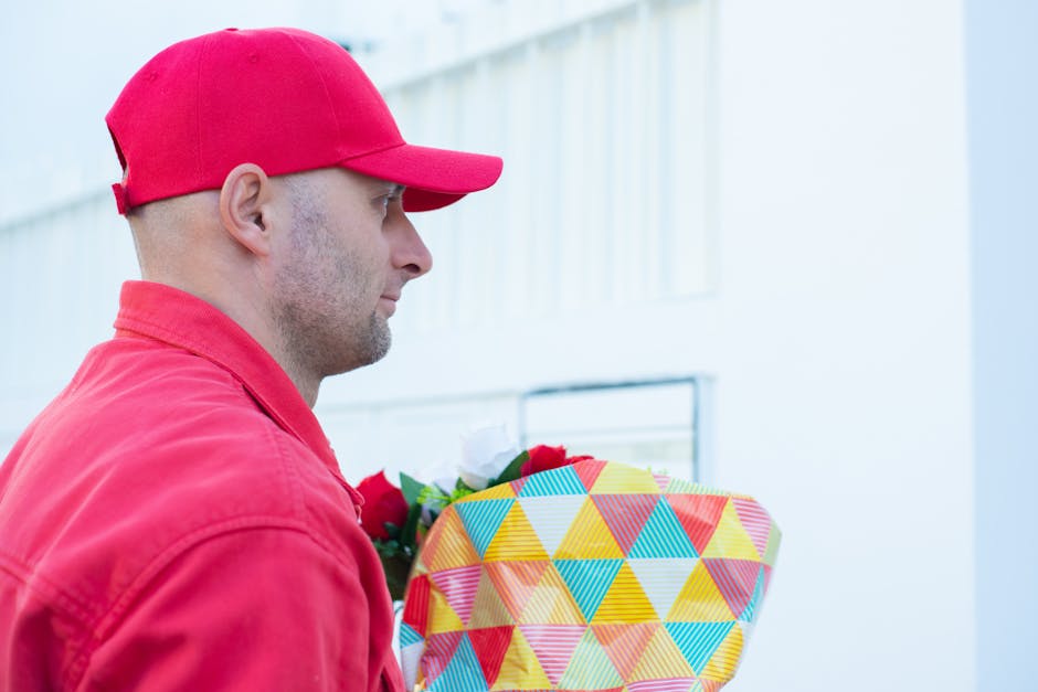 A delivery person dressed in a red cap and red jacket holds a bouquet of fresh flowers wrapped in decorative paper with a geometric pattern, featuring vibrant colors such as yellow, red, blue, and ora