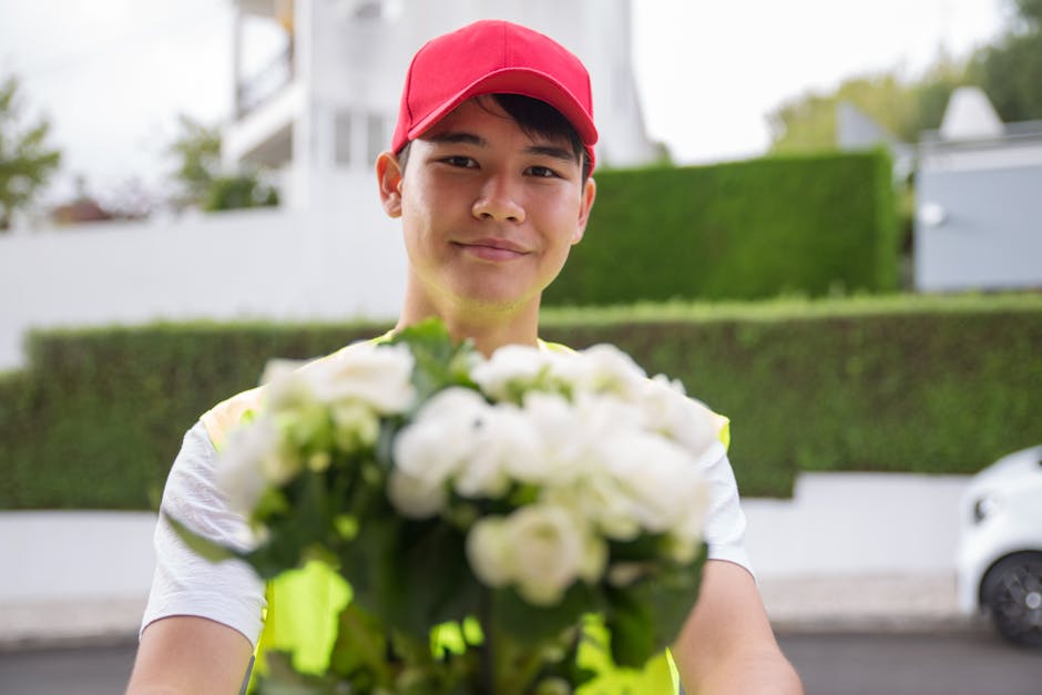A delivery person wearing a red cap and high-visibility vest holds out a bouquet of fresh white roses and greenery toward the camera. The bouquet is wrapped in clear cellophane and arranged with white