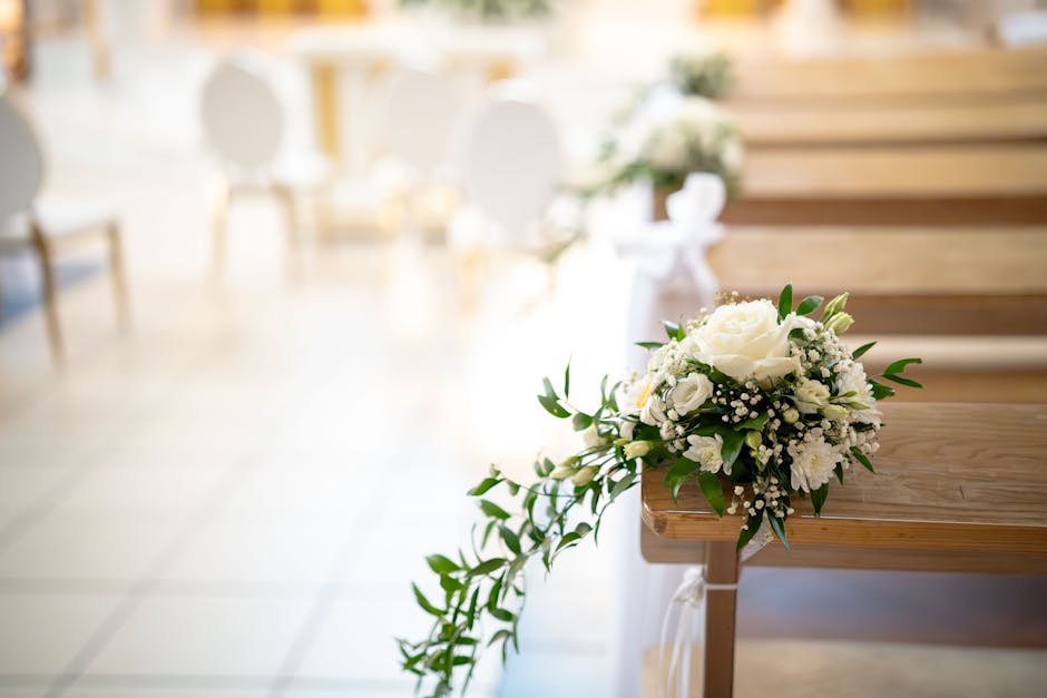 A close-up of a wedding bouquet resting on the edge of a wooden pew during a ceremony, featuring white roses, orchids, and baby's breath accented with green foliage. The bouquet is gathered with a sat