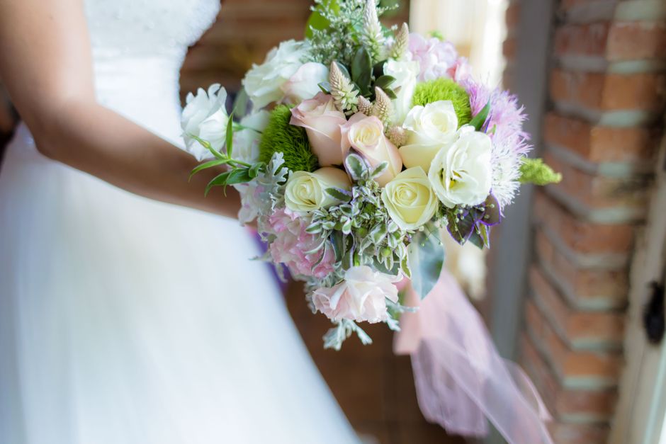A close-up of a bridal bouquet featuring white and pale pink roses, light purple and green hydrangeas, and sprigs of greenery, arranged in a rounded, natural style. The bouquet is wrapped in soft pink