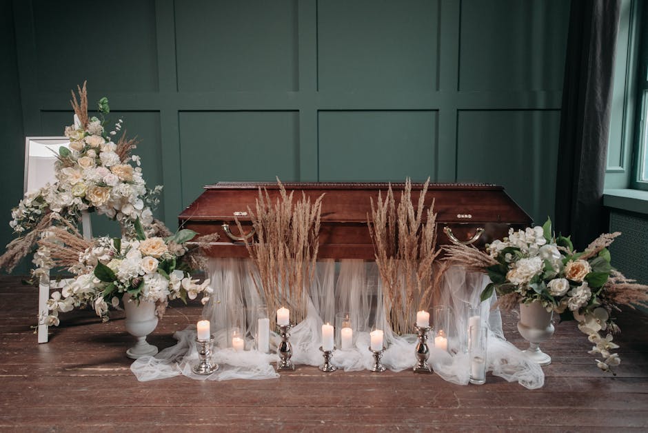 A floral arrangement placed on top of a wooden casket, featuring an elegant display of white roses, hydrangeas, and assorted greenery, accented with tall, beige dried pampas grass. The flowers are arr