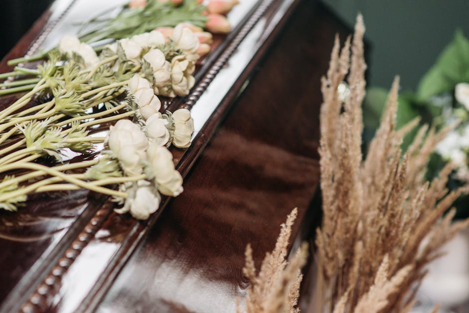 A close-up of a delicate floral arrangement placed on a polished wooden surface, featuring a linear bouquet of white roses with slightly ruffled petals, green stems, and buds, arranged neatly along a