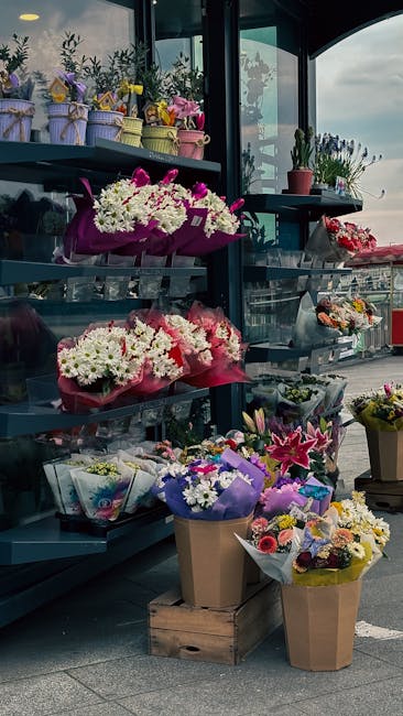 An outdoor display of various floral arrangements on metal shelves, featuring a selection of fresh flowers including white, pink, and purple blooms such as lilies, chrysanthemums, and roses. The arran