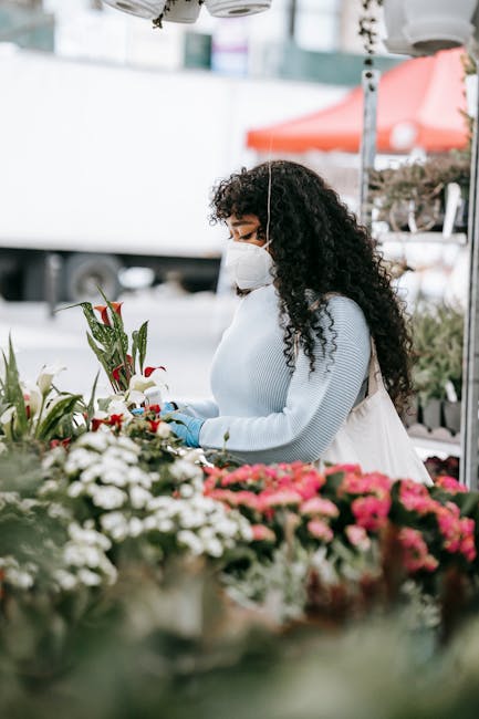 A woman with dark, curly hair wearing a white face mask and light blue gloves is arranging a floral bouquet at an outdoor flower market. She is dressed in a light blue sweater and a skirt, standing be