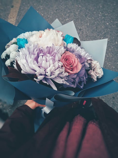 A person holding a beautifully arranged bouquet of fresh flowers from flowerdeliverylittleilford.co.uk, featuring a large white chrysanthemum, soft pink roses, purple statice, and small blue accents,