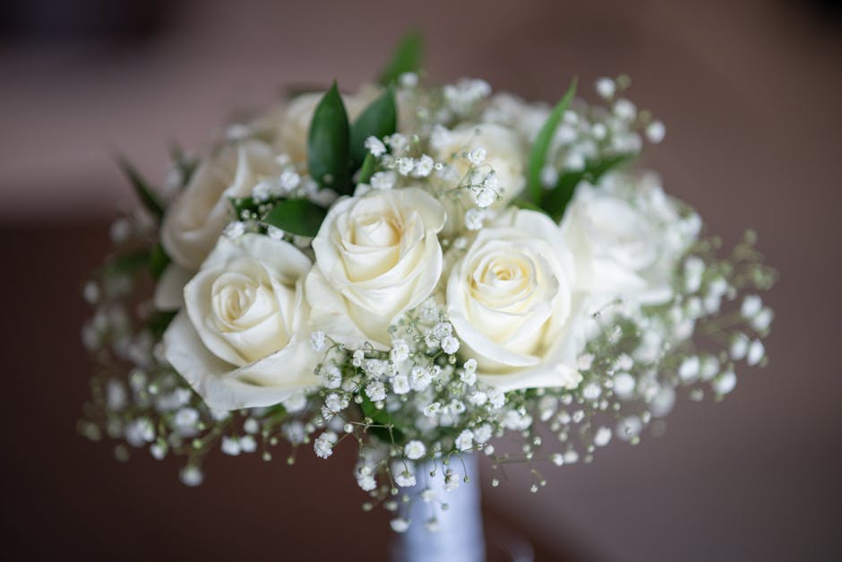 A bouquet arrangement of fresh white roses with tightly curled petals, complemented by delicate sprigs of baby’s breath and green foliage, is displayed in a glass vase. The bouquet is placed on a da