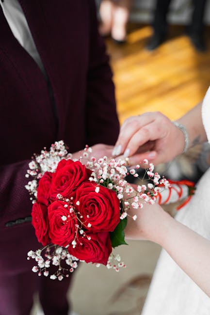 A bouquet of vibrant red roses, accented with small white baby's breath flowers, is being held by a person dressed in a white sleeve, likely during a wedding or floral gift presentation. The arrangeme