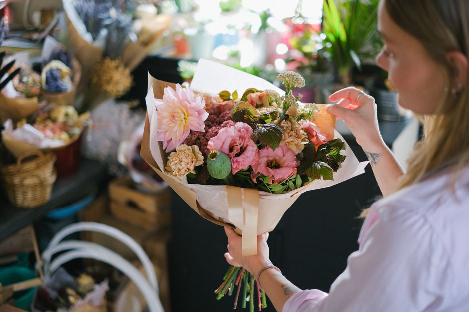 A woman holding a bouquet of fresh flowers in a floral shop, with a focus on pink and cream blooms including dahlias, roses, and lisianthus, wrapped in white and beige paper with a cream ribbon. The b