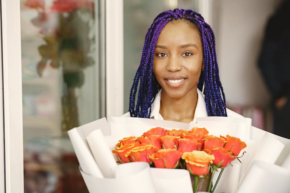 A young woman with dark skin and vibrant purple braided hair holds a large bouquet of freshly cut coral and yellow roses wrapped in white paper. She is smiling gently, standing indoors near what appea